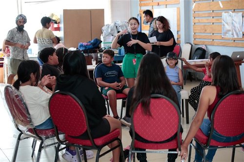 Kids and adults seated in a circle during a group activity in a room with chairs and tables.